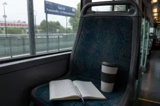Open notebook and coffee cup on bus seat near window, early morning light, rain-speckled glass, Val-d'Oise signage faintly visible outside.