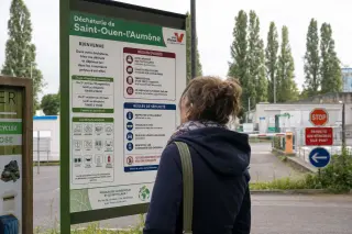 A woman reads recycling instructions at a Val-d'Oise recycling center entrance on a bright day, with visible local signage and natural surroundings.
