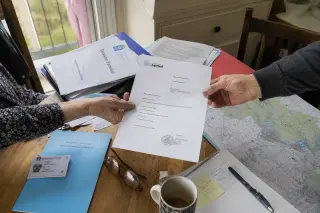 Overhead view of a wooden table with folders, letters, a local map, coffee mug, and two hands exchanging a document in natural morning light.