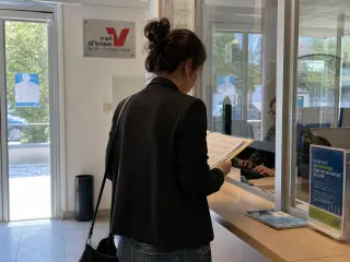 Resident in a municipal office in Val-d'Oise preparing documents at a service counter with clerk behind glass, in natural light.