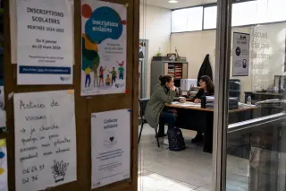 Close-up of a school enrollment bulletin board in a Val-d'Oise municipal office, with a young parent conversing with a staff member in the background, natural daylight illuminating the space.