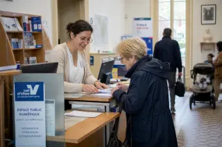 An elderly woman speaks with a municipal clerk at a mairie counter in the Val-d'Oise, surrounded by papers and softly lit by daylight through large windows.