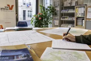 Resident's hand filling paperwork on a cluttered table with blueprints and notes in a sunny Val-d'Oise town hall office