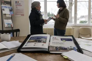 Social service brochures and notes on a table in a Val-d'Oise office, with two adults discussing in the background near a bright window.